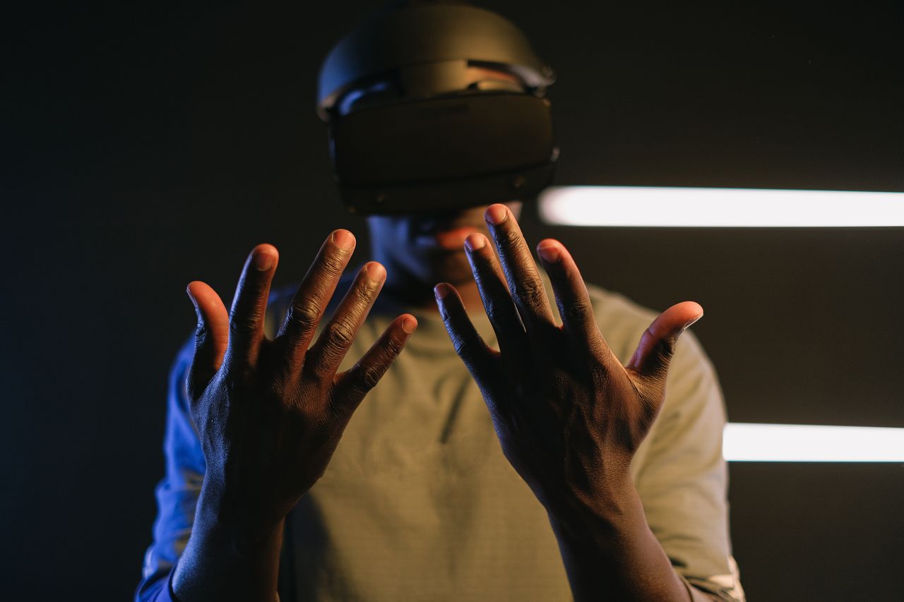 A Black man in a technology setting is using VR goggles with his arms extended.